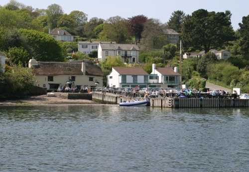 A view of Pandora Inn, in Falmouth, from the sea. The pontoon is busy with pub dwellers and a boat in moored up to the side.