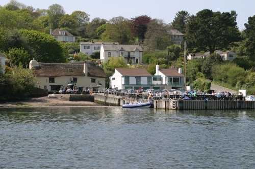 A view of Pandora Inn, in Falmouth, from the sea. The pontoon is busy with pub dwellers and a boat in moored up to the side.