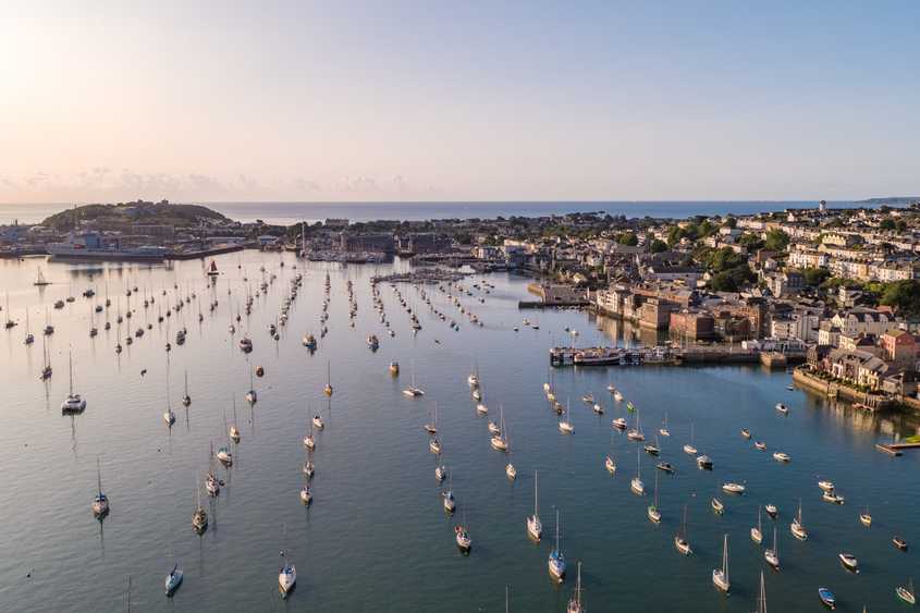 An aerial image of Falmouth Harbour at sunset, light bounces off the water and there are many boats moored on the harbour. The view stretches from Pendennis Castle to the Packet Quays.