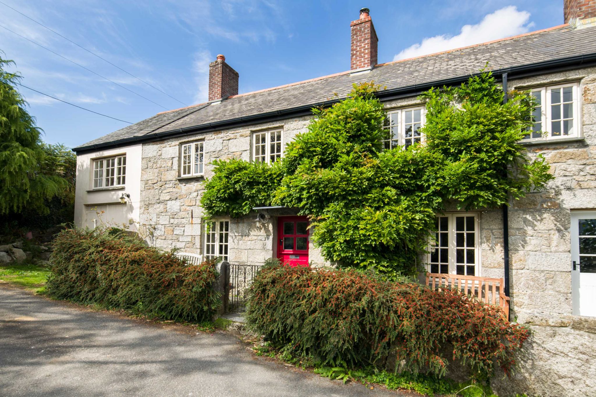 Pretty frontage of Pumpkin Cottage, a stone cottage with a red door covered in wisteria leaves and a blue sky behind